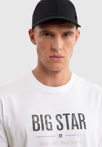 Giovane uomo che indossa un berretto nero e una maglietta bianca con la scritta "BIG STAR" sul davanti, che guarda dritto davanti a sé con un'espressione neutra.