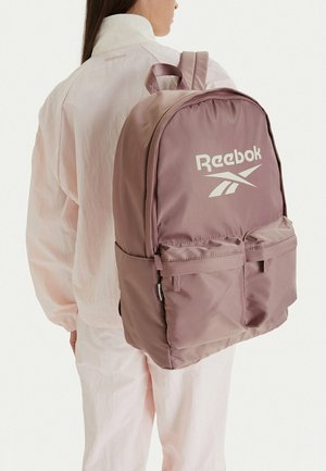 Persona con una mochila Reebok rosa polvoriento con bolsillos frontales, vestida con chaqueta y pantalones rosa claro, vista desde atrás.