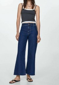 Jean évasé bleu marine en denim, doté d'une taille haute, d'un ourlet brut et d'un design à cinq poches, associé à un débardeur gris et des tongs.