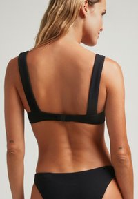 Top de bikini negro con canalé y tirantes anchos, que cuenta con un cierre trasero seguro. El diseño es minimalista, con una textura suave y acentos simples.