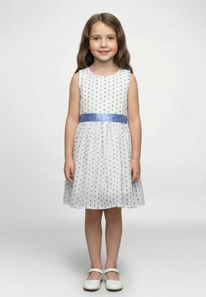 Vestito bianco senza maniche con motivo a pois blu, vita arricciata e nastro di raso blu. Abbinato a scarpe bianche, su uno sfondo neutro.