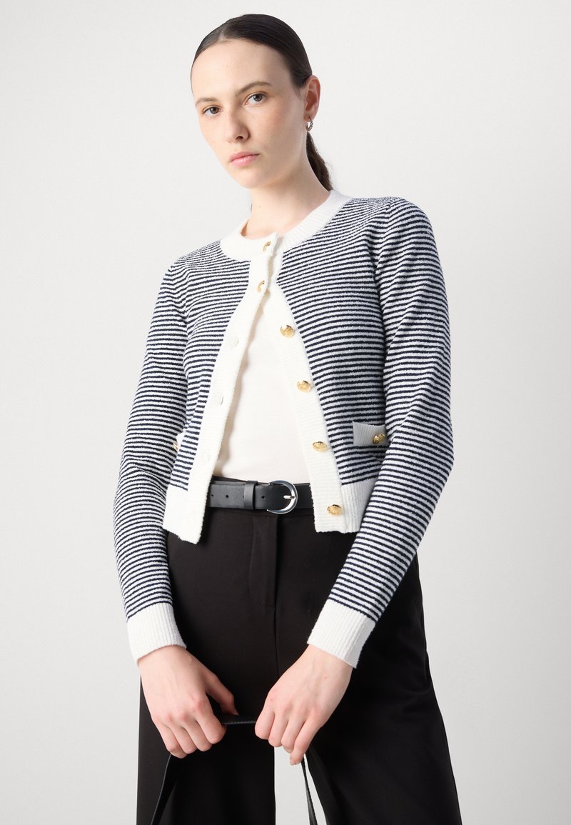 Cardigan rayé en bleu marine et blanc, avec des boutons dorés, un col rond et des poignets côtelés. Associé à un haut crème et une ceinture noire.