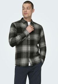 Grijs en zwart geruite flanellen shirt met knoopsluiting, lange mouwen en een kraag. Model past het shirt aan, gecombineerd met een marineblauwe broek.
