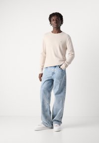 Light beige knitted sweater with long sleeves, paired with loose-fitting light blue denim jeans and white sneakers. Subtle logo detail on sweater.