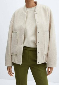 Beige wollen jasje met drukknoop over een bijpassende trui, gecombineerd met hooggetailleerde olijfgroene pantalon.