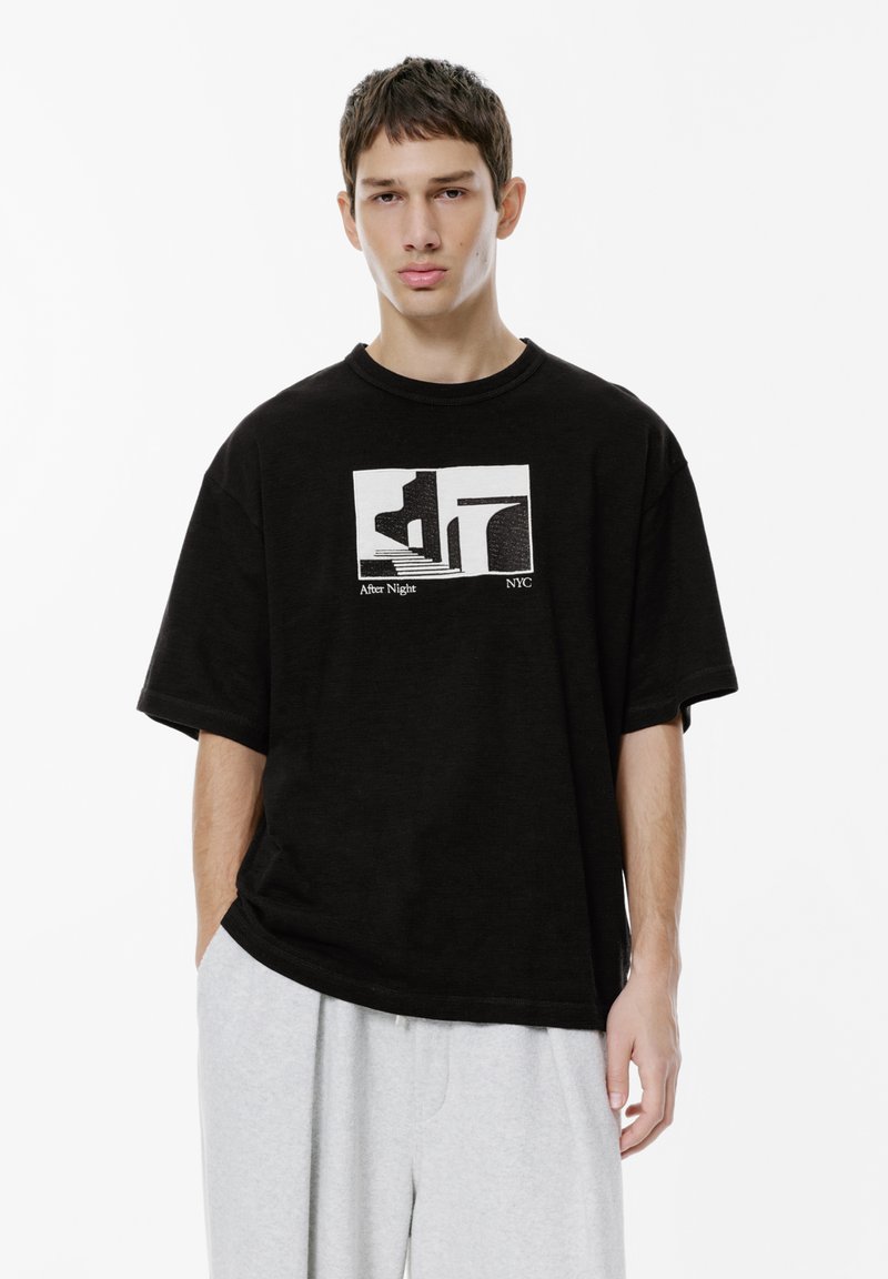 Ung mand iført en løs sort T-shirt med abstrakt hvid grafik og teksten "After Night NYC", parret med lysegrå bukser på hvid baggrund.