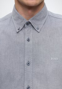 Gros plan d'une personne portant une chemise grise boutonnée avec un col boutonné et "BOSS" brodé sur la poitrine.