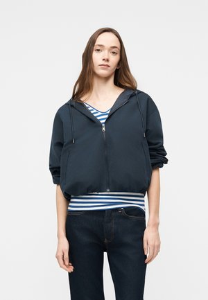 Ung kvinde iført en navyblå lynlåshættetrøje over en blå- og hvidstribet skjorte med mørk denimjeans, stående foran en ensfarvet baggrund.