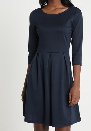 Kobieta ubrana w granatową sukienkę do kolan z rękawami do łokcia i dopasowaną talią, z plisami na spódnicy.