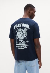 Tamsiai mėlynas medvilninis marškinėliai su žaisminga grafika ant nugaros, užrašas "ŽAISK ŠALTĄ" ir stilizuotas personažas su krepšiniu ir riedlente.