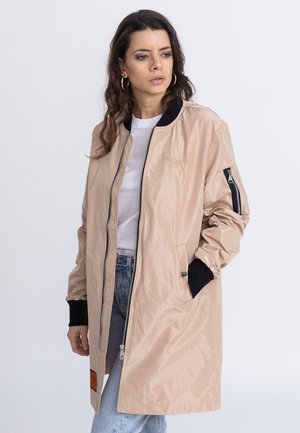 Veste bomber beige avec une fermeture éclair, col et poignets noirs, poche à glissière sur la manche et un patch sur l'ourlet inférieur. Texture lisse.