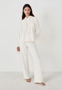 Frau steht barfuß in einem entspannten cremfarbenen Hoodie und weiten Hosen vor einem schlichten hellen Hintergrund.