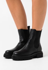 Bottines en cuir noir à semelles épaisses portées sur des jambes nues sur fond blanc.