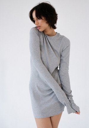 Kobieta o krótkich ciemnych włosach, ubrana w dopasowaną, jasnoszarą sukienkę z długim rękawem, stojąca na tle jednolitego, jasnego tła.