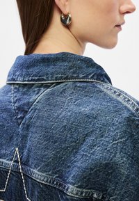 Τζάκετ από τζίν με μύτη κολάρο, σκούρο μπλε χρώμα και άσπρες ραφές που σχηματίζουν ένα μοτίβο αστεριού στην πλάτη. Μεγάλο κυκλικό σκουλαρίκι σε χρυσό και μαύρο χρώμα.