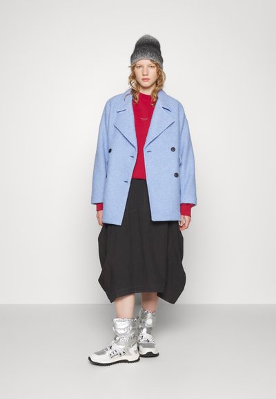 Manteau bleu clair à double boutonnage, pull rouge, jupe noire et bottes argentées avec des poignets froncés. Textures simples, couleurs variées et conceptions superposées.