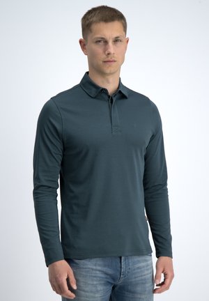 Jonge man met kort haar, gekleed in een donker petrolkleurige lange mouw polo en lichtblauwe spijkerbroek, staand voor een effen witte achtergrond.