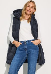 Gilet matelassé marine avec capuche, doté d'une fermeture éclair et de poches latérales. Assorti à une chemise blanche à manches longues et un jean bleu.