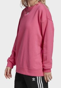 Rosa fleece sweatshirt med rund hals och sänkta axlar. Har vit Adidas-logotyp på bröstet och ribbade ärmslut. Matchas med svarta byxor.