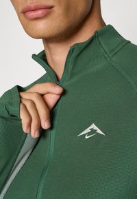Žalias zip-up sportinis džemperis su tekstūruotu audiniu, aukštu apykakle ir siuvinėtu logotipu. Yra lieso silueto ir daliniu užtrauktuku.