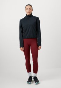 Črna športna jakna s četrtinskim zadrgo in teksturirano tkanino ter stranskim žepom, kombinirana z vinsko rdečimi pajkicami in črnimi supergami, z belimi nogavicami.