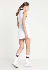 Robe de tennis sans manches blanche avec des rayures navy et un col, associée à une jupe évasée blanche et des chaussettes montantes blanches avec des rayures rouges et bleues.