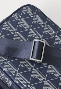 Borsa texture blu navy con motivo geometrico bianco, dotata di tracolla in tessuto e fibbia metallica. Logo "LACOSTE" esposto in modo prominente.