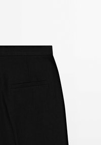 Pantalon noir sur mesure avec une ceinture plate et une seule poche passepoilée à l'arrière côté droit sur fond blanc.