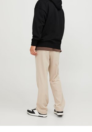 Personne portant un pantalon beige, des baskets noires et blanches, et un sweat à capuche noir par-dessus un t-shirt marron, debout de profil sur un fond uni.