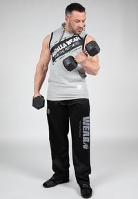 Grijze mouwloze hoodie met zwarte accenten, voorzien van tekst. Zwarte joggerbroek met "WEAR" in het wit gedrukt, terwijl hij dumbbells vasthoudt, neutrale achtergrond.