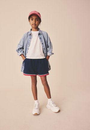 Ung pige står med hænderne i lommerne iført en lyserød kasket, denimskjorte, hvid t-shirt, marineblå shorts med røde kanter, hvide sokker og sneakers.