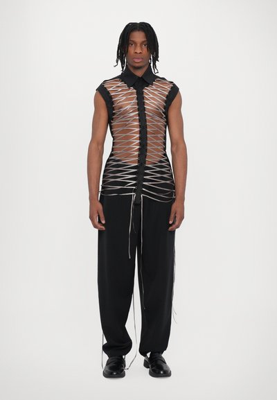 Haut transparent beige avec patte de boutonnage noire et design en tissage ouvert, associé à un pantalon noir ample et des chaussures noires.