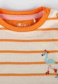 Gestreiftes T-Shirt in Creme und Orange, mit einem blauen gestickten Flamingo auf Rädern und roten Akzenten, Rundhalsausschnitt und Druckknöpfen.