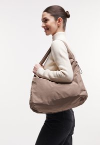 Grote beige schoudertas van zacht materiaal, met een ronde vorm en een enkele schouderband. Subtiele branding in reliëf aan de voorkant.