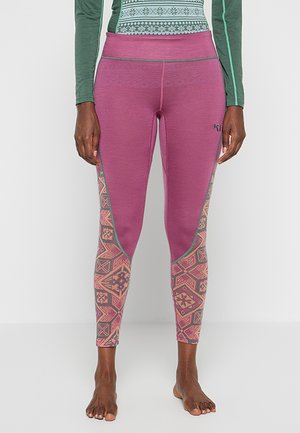 Frau trägt violette Leggings mit pinken und gemusterten geometrischen Designs an den unteren Beinen, kombiniert mit einem grünen, gemusterten Langarmshirt.