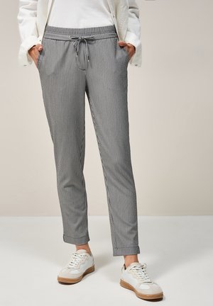 Osoba w czarno-białych pionowo paskowanych spodniach na sznurku, białych trampkach i białej kurtce, z rękami w kieszeniach.