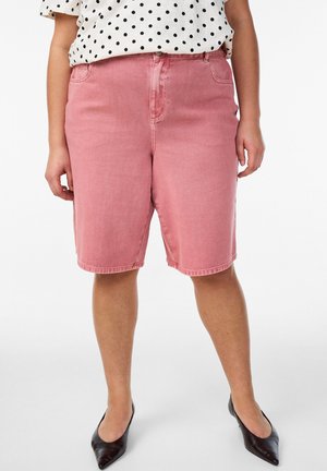 Naine, kellel on keskmise pikkusega roosad teksapüksid, mustad teravnurkse ninaga kingad ja valge pluus mustade täppidega, seisab ühevärvilise tausta ees.