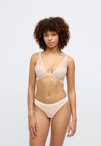 Bež set donjeg rublja koji se sastoji od prozirnog grudnjaka s mrežastim panelima i glatkim žicom, uparenog s odgovarajućim tangama koje prikazuju logo.