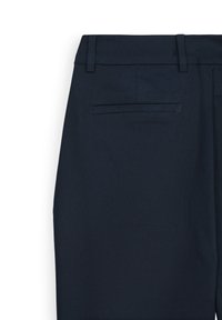 Dunkelblaue Anzughosen mit Gürtelschlaufen und einer einzigen Paspeltasche auf der Rückseite rechts vor einem weißen Hintergrund.