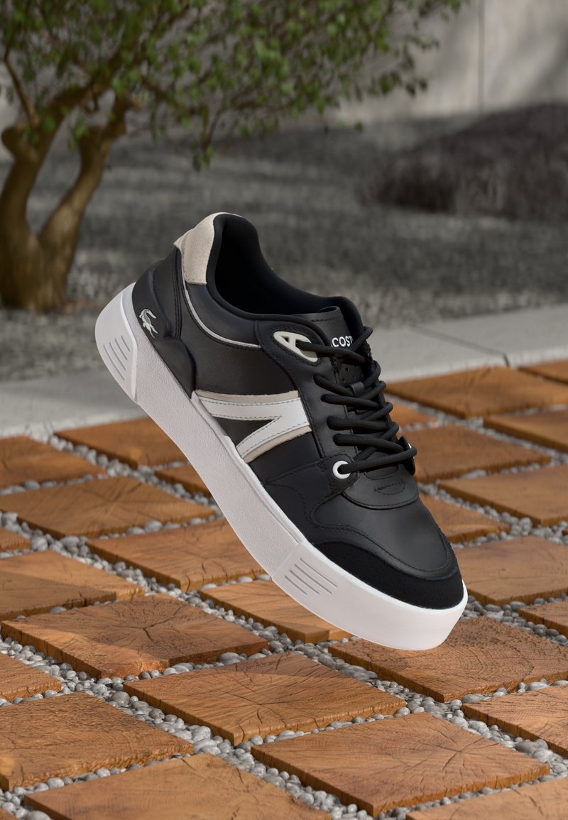 Baskets en cuir noir avec des accents blancs, présentant un dessus texturé, une semelle blanche et des lacets noirs, flottant au-dessus d'une surface à motif en pierre.