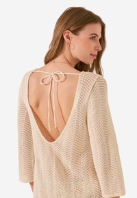 Top lavorato a maglia di colore crema con scollo a V profondo sul retro e chiusura con laccetto, maniche ampie e un motivo testurizzato su tutta la superficie.