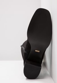 Botte en daim noir à talon épais posée sur une surface blanche, montrant la semelle lisse avec une petite étiquette de marque dorée près de la voûte plantaire.