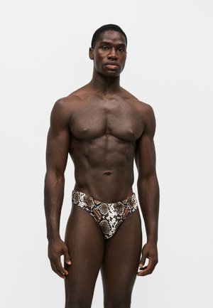 Homme musclé à la peau foncée portant un slip de bain marron et blanc à motif serpent, debout devant un fond clair uni.