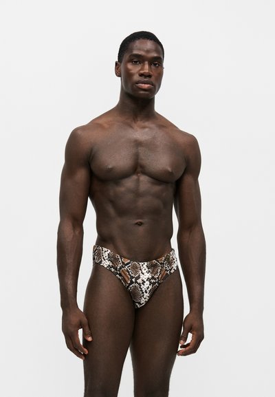 Homme musclé à la peau foncée portant un slip de bain marron et blanc à motif serpent, debout devant un fond clair uni.