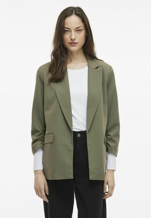 Olivově zelený blazer se strukturovaným designem, přední kapsou a ohrnutými rukávy, nošený přes bílý top a černé kalhoty.