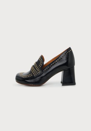 Mocassin à talon en cuir verni noir avec un talon bloc et des clous dorés le long des bords et une bride sur le dessus.