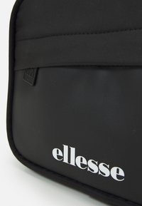 Černá nylonová taška s hladkým povrchem, s bílým logem "ellesse" a malým kovovým štítkem. Obdélníkový tvar se zapínáním na zip.