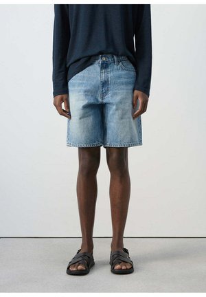 Persona que lleva pantalones cortos de mezclilla azul claro, camiseta negra de manga larga y sandalias negras abiertas, de pie sobre un suelo gris.