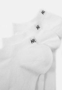 Weiße Knöchelsocken mit gerippten Bündchen, die jeweils mit einem schwarzen Markenlogo versehen sind. Sie haben eine glatte Textur und ein standardmäßiges sportliches Design.