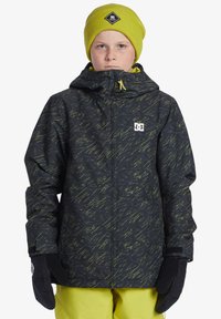 Svart och gula mönstrade snöjacka med huva, framdragkedja och synlig DC-logga. Bärs med en matchande gul stickad mössa och svarta handskar.